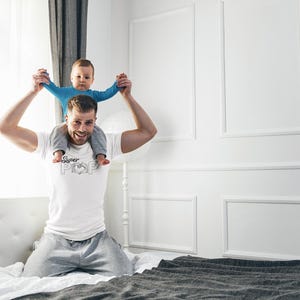 May include: A man wearing a white t-shirt with the text "Super Pop" and a heart graphic, holding a baby on his shoulders. The baby is wearing a blue long-sleeved shirt. They are in a bedroom with white walls and a bed.