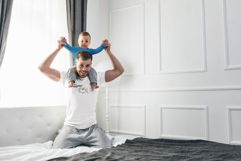 May include: A man wearing a white t-shirt with the text "Super Pop" and a heart graphic, holding a baby on his shoulders. The baby is wearing a blue long-sleeved shirt. They are in a bedroom with white walls and a bed.