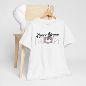 Peut inclure: Un t-shirt blanc &agrave; manches courtes avec le texte noir Super Grand MOM, o&ugrave; la lettre O est remplac&eacute;e par deux mains formant un c&oelig;ur avec un motif floral color&eacute;. Une casquette de baseball blanche unie est suspendue sur la chaise en bois.