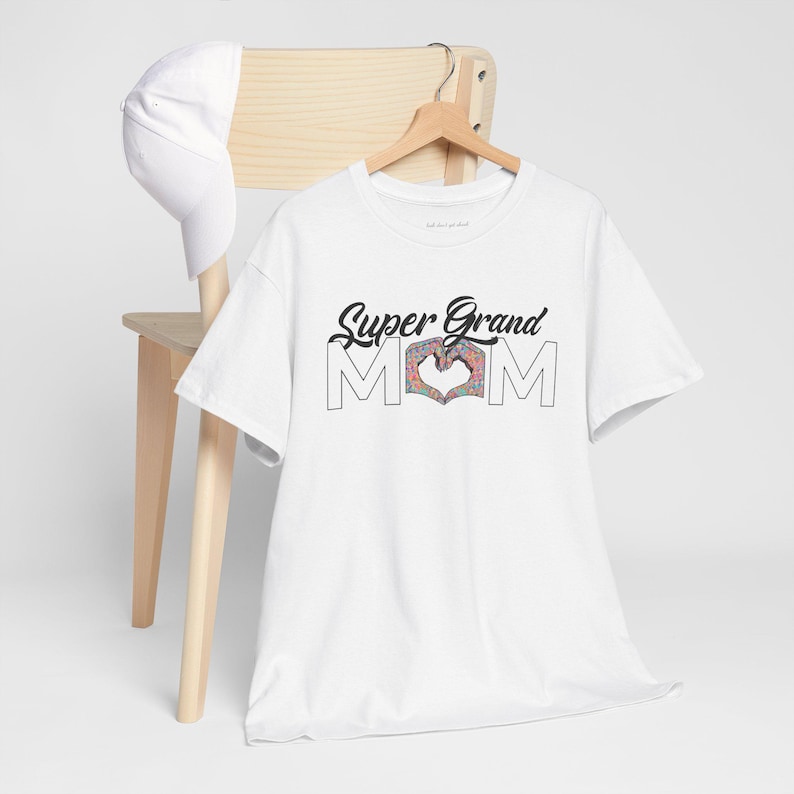 Peut inclure: Un t-shirt blanc &agrave; manches courtes avec le texte noir Super Grand MOM, o&ugrave; la lettre O est remplac&eacute;e par deux mains formant un c&oelig;ur avec un motif floral color&eacute;. Une casquette de baseball blanche unie est suspendue sur la chaise en bois.
