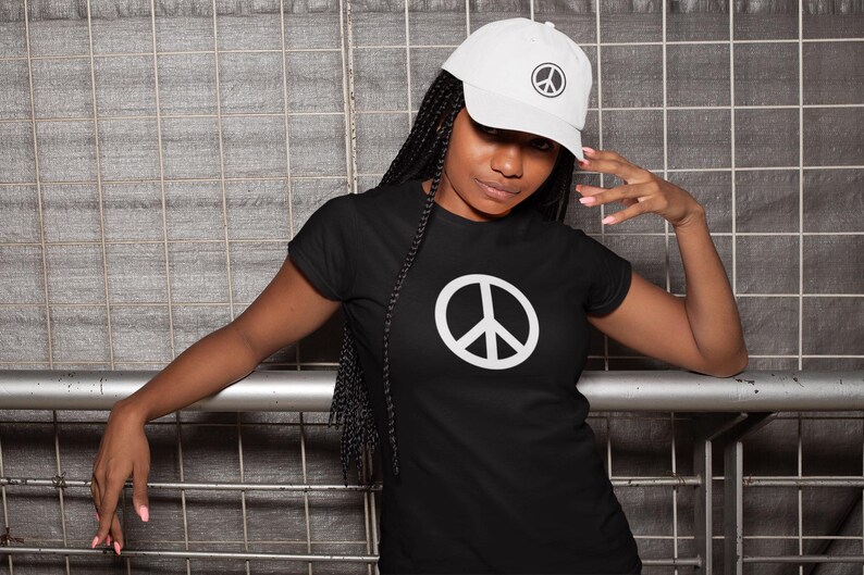 May include: Black t-shirt and white baseball cap, both featuring a white peace symbol. The t-shirt has a large peace sign in the center. The cap has a smaller peace symbol on the front. The person is leaning against a metal structure.