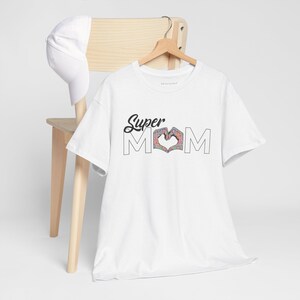 May include: A white short-sleeved t-shirt featuring the black text Super MOM, where the letter O is replaced by two hands forming a heart shape with a colorful floral pattern. A plain white baseball cap hangs on the wooden hanger with the shirt.
