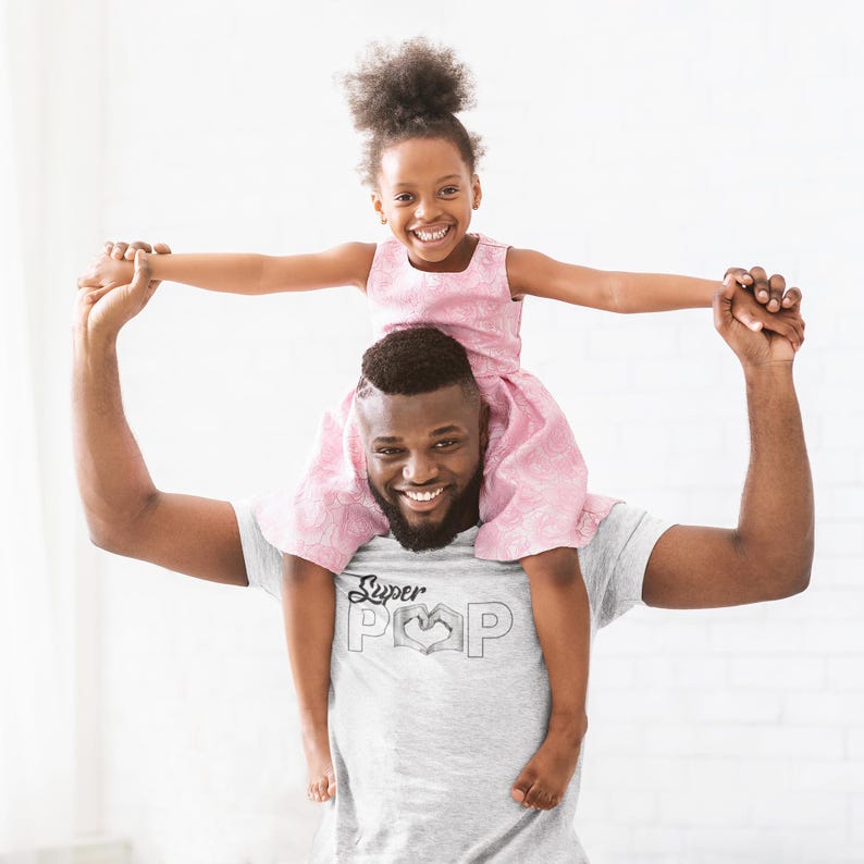 May include: A smiling man wearing a gray t-shirt with the words "Super Pop" and a heart-shaped hand symbol, holding a smiling girl wearing a pink dress on his shoulders. The background is a plain white wall.