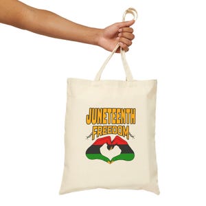 May include: Cream-colored tote bag with the words "Juneteenth Freedom" in yellow and black. The design includes a heart shape formed by hands in the Pan-African colors of red, black, and green. The bag is held by a person.