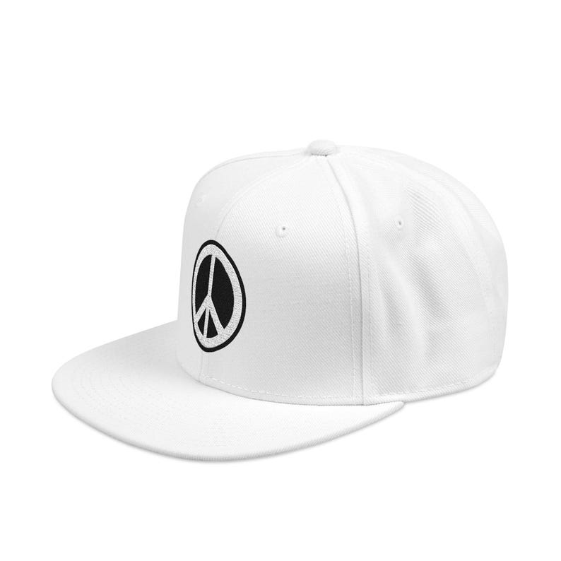 May include: A white baseball cap featuring a black circular peace sign emblem embroidered on the front panel. The cap has a structured crown, a flat brim, and a button at the top center, constructed from a solid white fabric material.