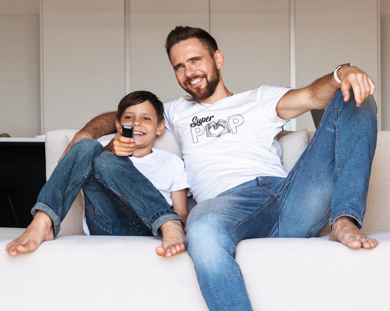 May include: A father and son are seated on a cream-colored sofa. Both are wearing white t-shirts and blue jeans. The father's shirt has the words "Super Pop" printed on it. The son is holding a remote control. They are smiling at the camera.