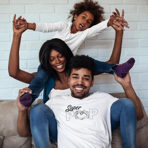 May include: A family of three is smiling and posing for a photo. The father is wearing a white t-shirt with the words "Super POP" in a heart shape. The mother and daughter are on top of the father's shoulders.
