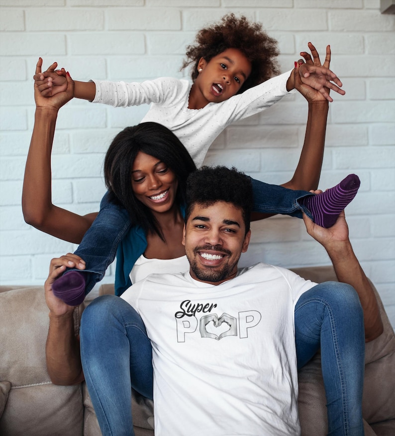 May include: A family of three is smiling and posing for a photo. The father is wearing a white t-shirt with the words "Super POP" in a heart shape. The mother and daughter are on top of the father's shoulders.
