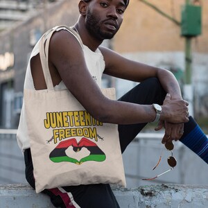 May include: Beige tote bag with the words "Juneteenth Freedom" in yellow and a heart-shaped design in red, black, and green. The bag has a shoulder strap and is being held by a person.