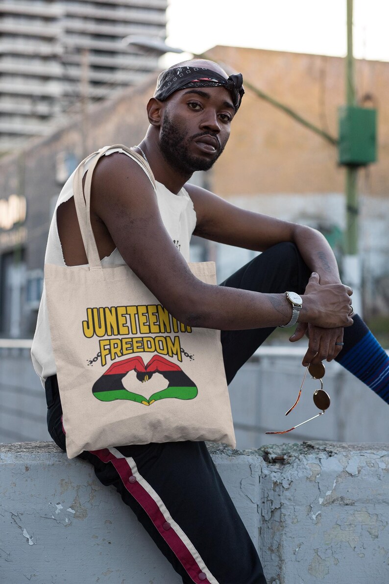 May include: Beige tote bag with the words "Juneteenth Freedom" in yellow and a heart-shaped design in red, black, and green. The bag has a shoulder strap and is being held by a person.