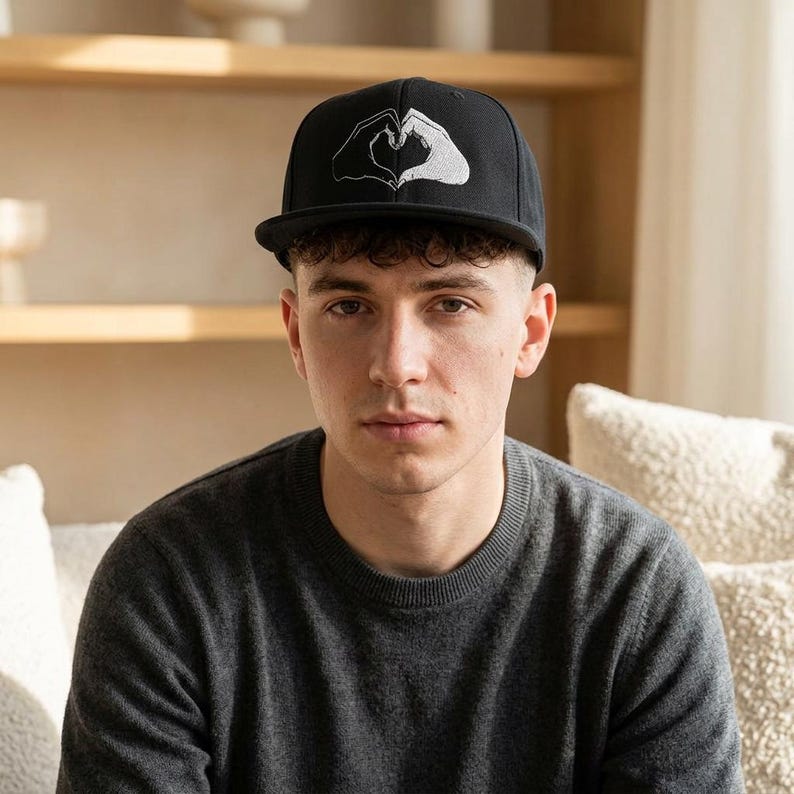 May include: A black baseball cap featuring a white embroidered graphic of two hands forming a heart shape on the front panel. The cap has a structured crown, a flat brim, and a classic six-panel design with a button on the top center.
