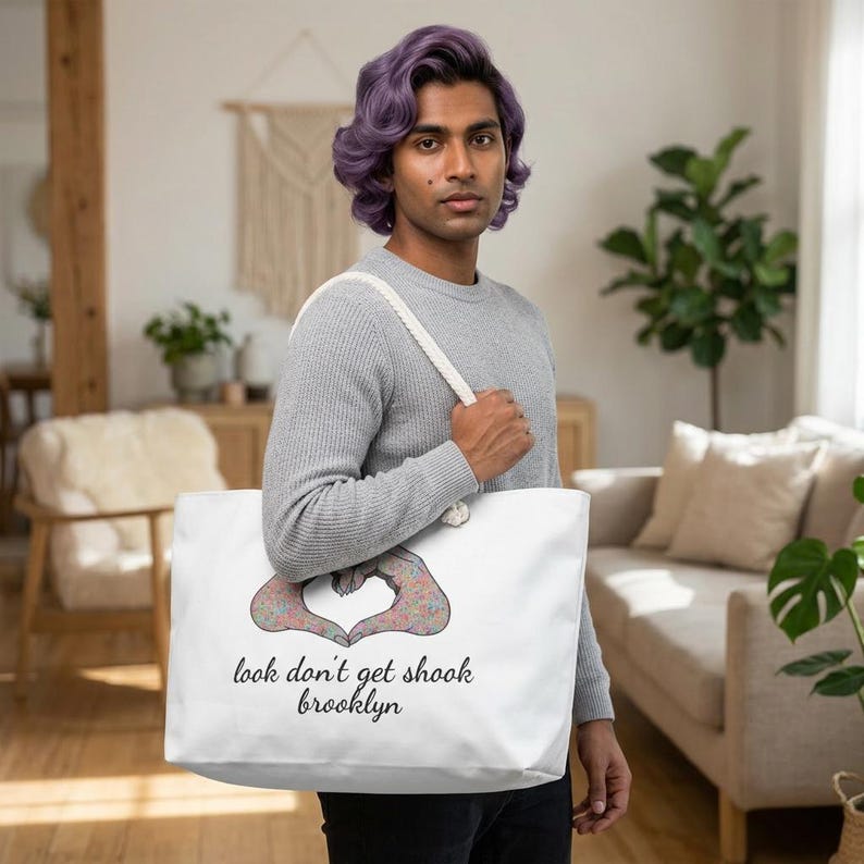 May include: White tote bag with a floral heart design and the text "look don't get shook brooklyn". The bag has a cream-colored strap and is held by a person wearing a gray sweater.