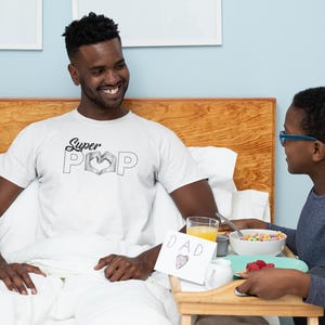 May include: A man wearing a white t-shirt with the text "Super Pop" and a heart design, sitting in bed. A child is bringing a breakfast tray with cereal, juice, and a card that says "DAD" with a heart.