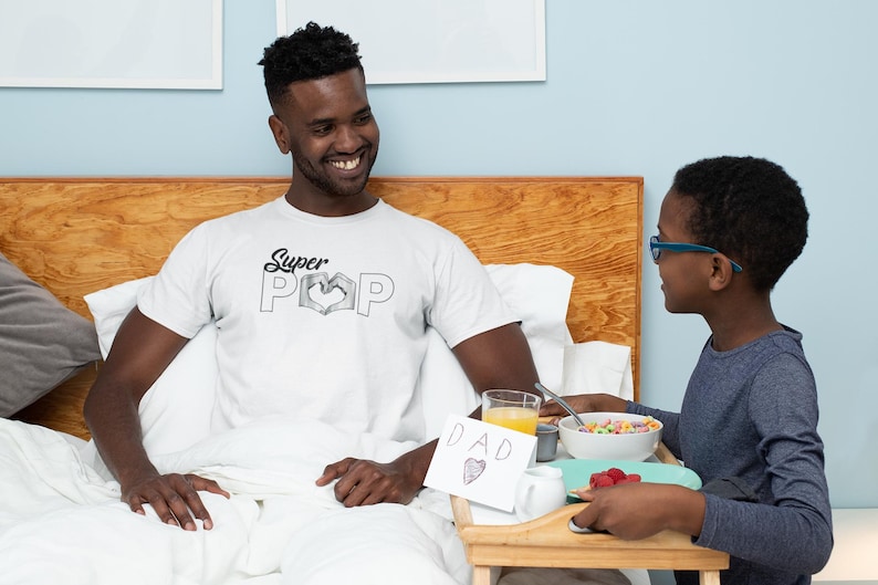 May include: A man wearing a white t-shirt with the text "Super Pop" and a heart design, sitting in bed. A child is bringing a breakfast tray with cereal, juice, and a card that says "DAD" with a heart.