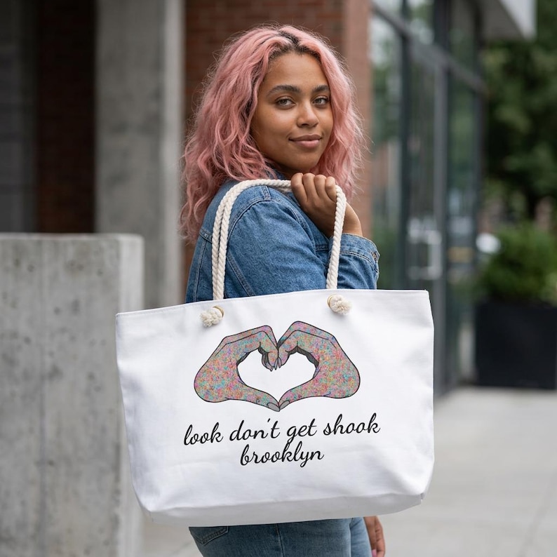 May include: White tote bag with rope handles featuring a heart made of hands with colorful patterns. The text "look don't get shook brooklyn" is printed below the heart design. The bag is held by a person.