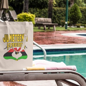 May include: A beige tote bag with the words "Juneteenth Freedom" in yellow and black lettering. The bag features a heart-shaped design in red, black, and green. The bag is resting on a lounge chair near a pool.