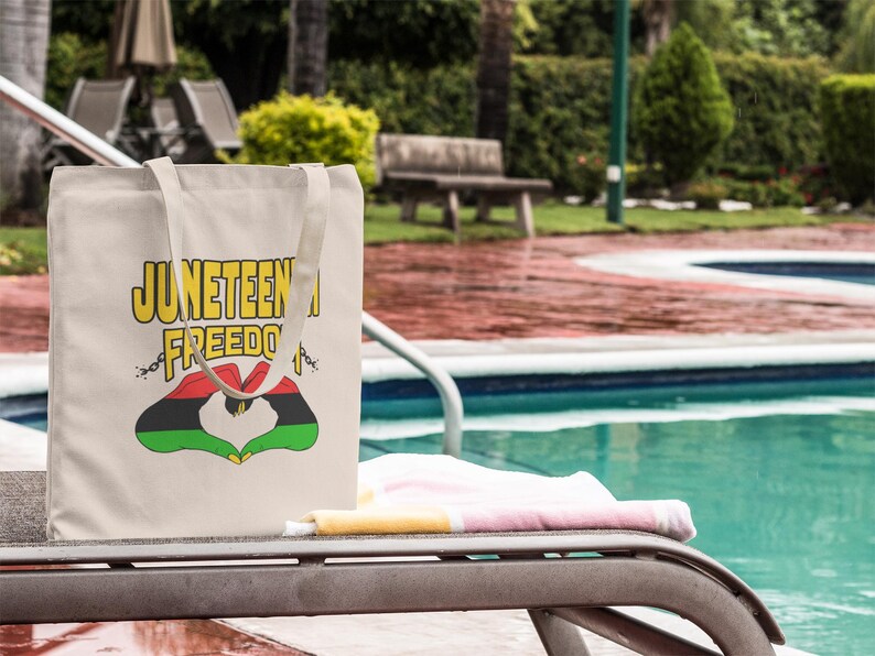 May include: A beige tote bag with the words "Juneteenth Freedom" in yellow and black lettering. The bag features a heart-shaped design in red, black, and green. The bag is resting on a lounge chair near a pool.