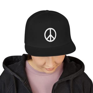 May include: A black baseball cap featuring a white embroidered peace sign symbol centered on the front panel. The cap has a structured crown and a flat brim, constructed from a solid black fabric with a clean, minimalist design aesthetic.