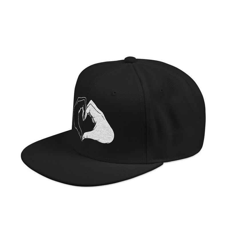 May include: A black snapback cap featuring a white embroidered graphic on the front panel. The design depicts two hands positioned together to form a heart shape, with detailed line work visible in the embroidery against the solid black fabric.