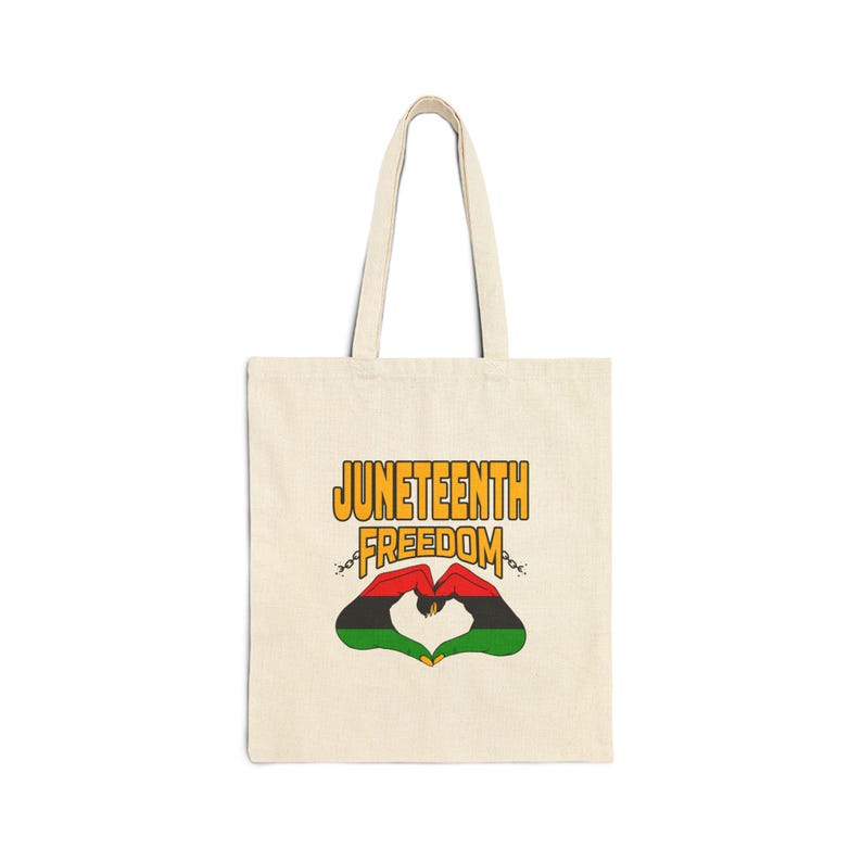 May include: Cream-colored tote bag with the words "Juneteenth Freedom" in yellow and black. The design includes a heart shape formed by red and green hands, with chains on either side.