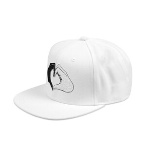 May include: A white snapback cap featuring a black and white graphic illustration on the front panel. The graphic depicts two hands positioned together to form a heart shape, with one hand rendered in solid black and the other in line art style.