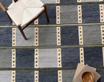 May include: A wooden chair with a woven seat sits on a rug featuring a grid pattern of dark blue and light blue rectangles, separated by vertical cream-colored bands with small brown and green squares, alongside a stack of books and a wooden bowl.