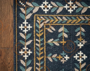 May include: Close-up of a navy blue rug with a detailed floral pattern. The design features white, teal, and tan leaf and cross motifs, framed by a gold border. The rug has a fringed edge, adding texture and detail to the design.