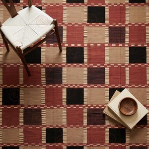 May include: A woven area rug featuring a repeating geometric pattern of large squares in terracotta red, black, and beige. The squares are separated by horizontal bands of small, dashed white lines. A wooden chair and a stack of books are present.