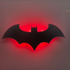 Puede incluir: Un logotipo negro de Batman con retroiluminación roja. El emblema en forma de murciélago está montado en una pared gris claro. Las alas del logotipo están extendidas, con una parte superior puntiaguda y una cabeza de murciélago central.
