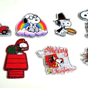 May include: A collection of Snoopy character pins in various designs. The pins feature Snoopy in different outfits and settings, including a firefighter, a rainbow, and a Dodgers baseball uniform. Some pins have glitter accents and the words "Merry Christmas".