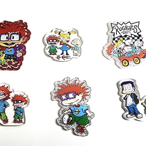 May include: A collection of six clear acrylic pins featuring characters from the animated series Rugrats. The pins showcase characters like Chuckie, Tommy, and Angelica, with glitter accents and vibrant colors.