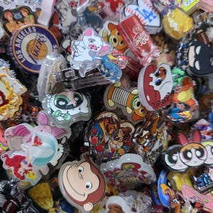 May include: A collection of colorful, cartoon-themed pins and charms. The assortment includes characters from popular animated movies, sports team logos, and various other designs. The pins are made of plastic and metal, with vibrant colors and detailed artwork.