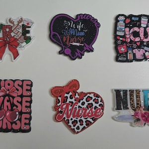 May include: A collection of six decorative pins with nurse-themed designs. The pins feature various shapes, including hearts and squares, and include text such as "Nurse" and "ICU". The color palette includes red, pink, black, and white.