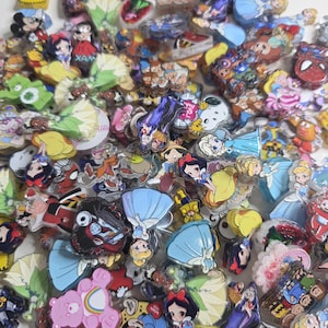 May include: A collection of colorful character-themed pins and charms. The assortment includes images of cartoon characters, princesses, and superheroes. The pins are made of various materials and feature bright colors and detailed designs.