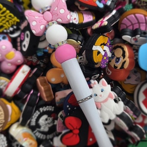 May include: A white pen with a pink and white top, surrounded by various colorful shoe charms. The pen has a silver band and tip. The charms include cartoon characters, bows, and other decorative elements.