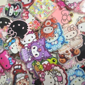 May include: A collection of colorful, glittery Hello Kitty character charms and accessories. The assortment includes various designs, such as Hello Kitty with a butterfly, in a mushroom hat, and other themed characters. The charms are in various colors, including pink, purple, and black.