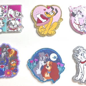 May include: A collection of colorful enamel pins featuring various Disney characters. Designs include Dalmatian puppies, Pluto, and characters from Lady and the Tramp. Each pin has glitter accents and a unique shape.