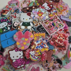 May include: A collection of colorful, glittery character pins and badges, including Hello Kitty, Mickey Mouse, and Disney princesses. The pins feature various shapes and designs, with Valentine's Day and other themes.