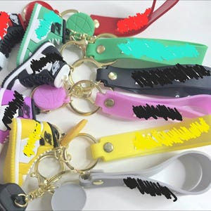 Può includere: Una collezione di portachiavi a forma di sneaker colorate. I portachiavi presentano design di scarpe in miniatura in vari colori, tra cui rosso, verde, nero, viola e giallo. Ogni portachiavi ha un anello color oro e un cinturino abbinato.