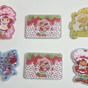 May include: A collection of colorful, glittery Strawberry Shortcake character-themed stickers and embellishments. The designs include the character in various poses, a bow, and rectangular designs with strawberry and glitter accents.