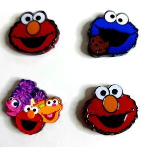 May include: Four colorful Sesame Street character pins. Includes Elmo, Cookie Monster with a cookie, and a group of characters. The pins have a glossy finish and are set against a white background.