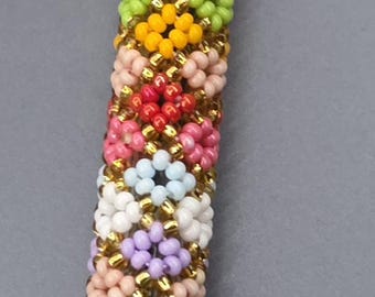 May include: A cylindrical pen covered in a vibrant, multi-colored beaded pattern. The design features small, circular flower-like motifs in shades of pink, yellow, blue, green, orange, purple, and white, separated by rows of metallic gold beads.