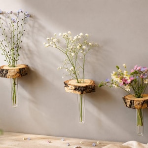 Könnte beinhalten: Drei Wandvasen mit Blumen. Jede Vase hat ein klares Glasrohr, das unter einer kreisförmigen, rustikalen Holzscheibe aufgehängt ist. Die Blumen umfassen blaue, weiße und rosa Blüten und schaffen eine natürliche, dekorative Präsentation.