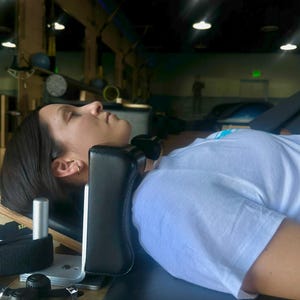 May include: A black padded headrest is attached to a wooden and metal exercise machine. A person is lying on the machine, wearing a white t-shirt. The machine is in a gym setting with overhead lights.