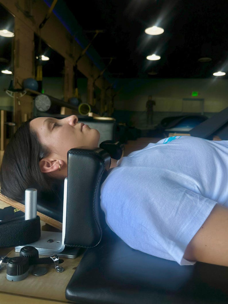May include: A black padded headrest is attached to a wooden and metal exercise machine. A person is lying on the machine, wearing a white t-shirt. The machine is in a gym setting with overhead lights.