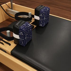 May include: A Pilates reformer machine with black leather padding and two padded blocks. The blocks are black with a navy blue patterned fabric and white straps. The machine is made of wood and metal.