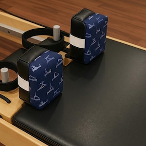 Puede incluir: Dos reposacabezas negros y azules para m&aacute;quinas de Pilates. Los reposacabezas tienen una tela azul con un patr&oacute;n blanco de poses de Pilates. Los reposacabezas son rectangulares con una banda blanca en el medio. La m&aacute;quina tiene correas negras y una superficie negra.