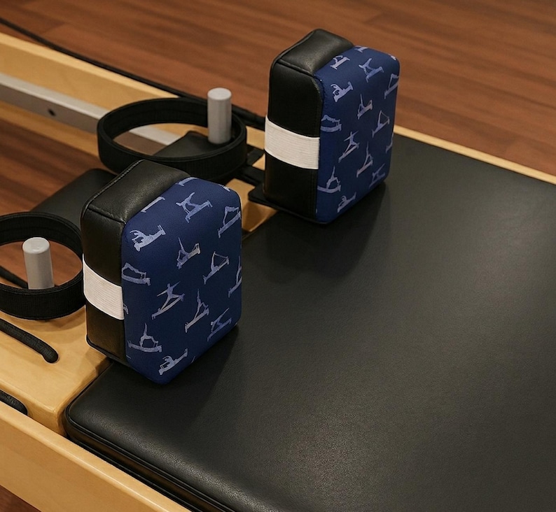 Puede incluir: Dos reposacabezas negros y azules para m&aacute;quinas de Pilates. Los reposacabezas tienen una tela azul con un patr&oacute;n blanco de poses de Pilates. Los reposacabezas son rectangulares con una banda blanca en el medio. La m&aacute;quina tiene correas negras y una superficie negra.