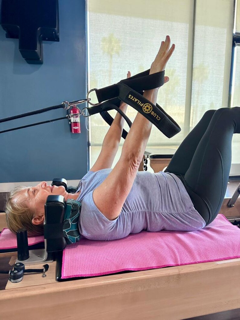 Puede incluir: Una mujer se ejercita en una m&aacute;quina de reformer Pilates. Lleva una camisa azul claro y leggings negros. La m&aacute;quina tiene correas negras con el texto "CLUB PILATES". Una esterilla rosa est&aacute; debajo de su espalda.
