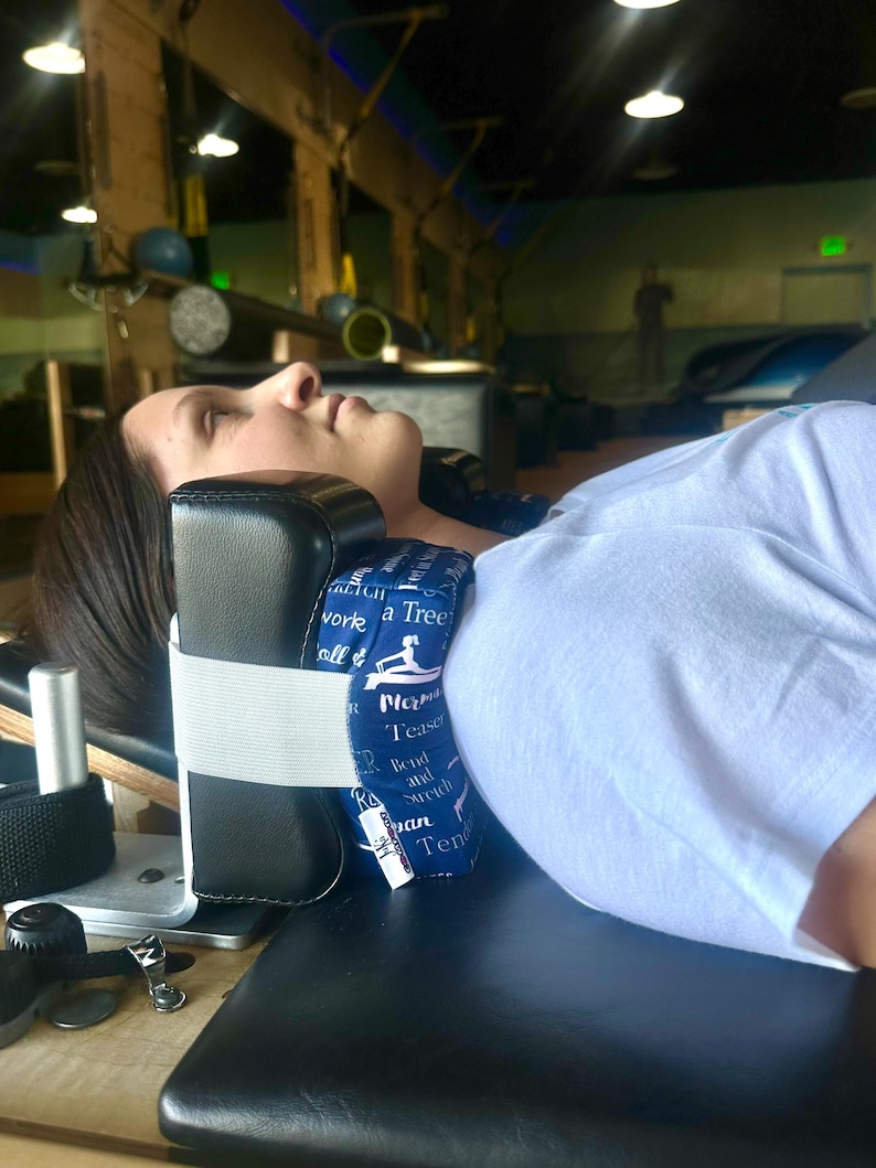 Puede incluir: Una almohada de soporte para el cuello en blanco y negro con una correa el&aacute;stica blanca. La almohada tiene tela azul con texto blanco y un gr&aacute;fico de yoga. La persona est&aacute; acostada sobre una superficie negra.